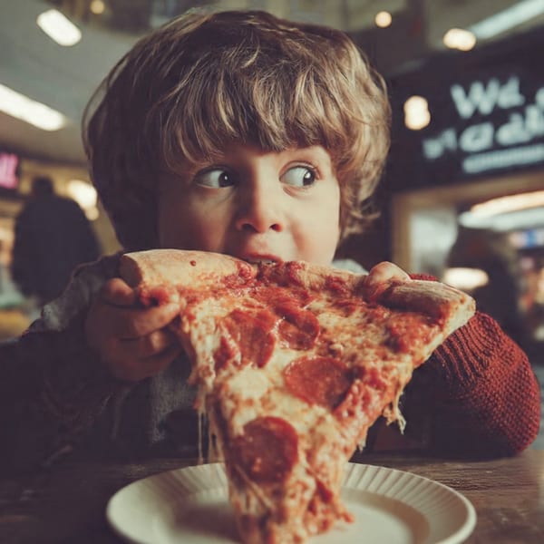 A young boy with curly hair takes a big bite of a pepperoni pizza slice at Italian Village Pizza.